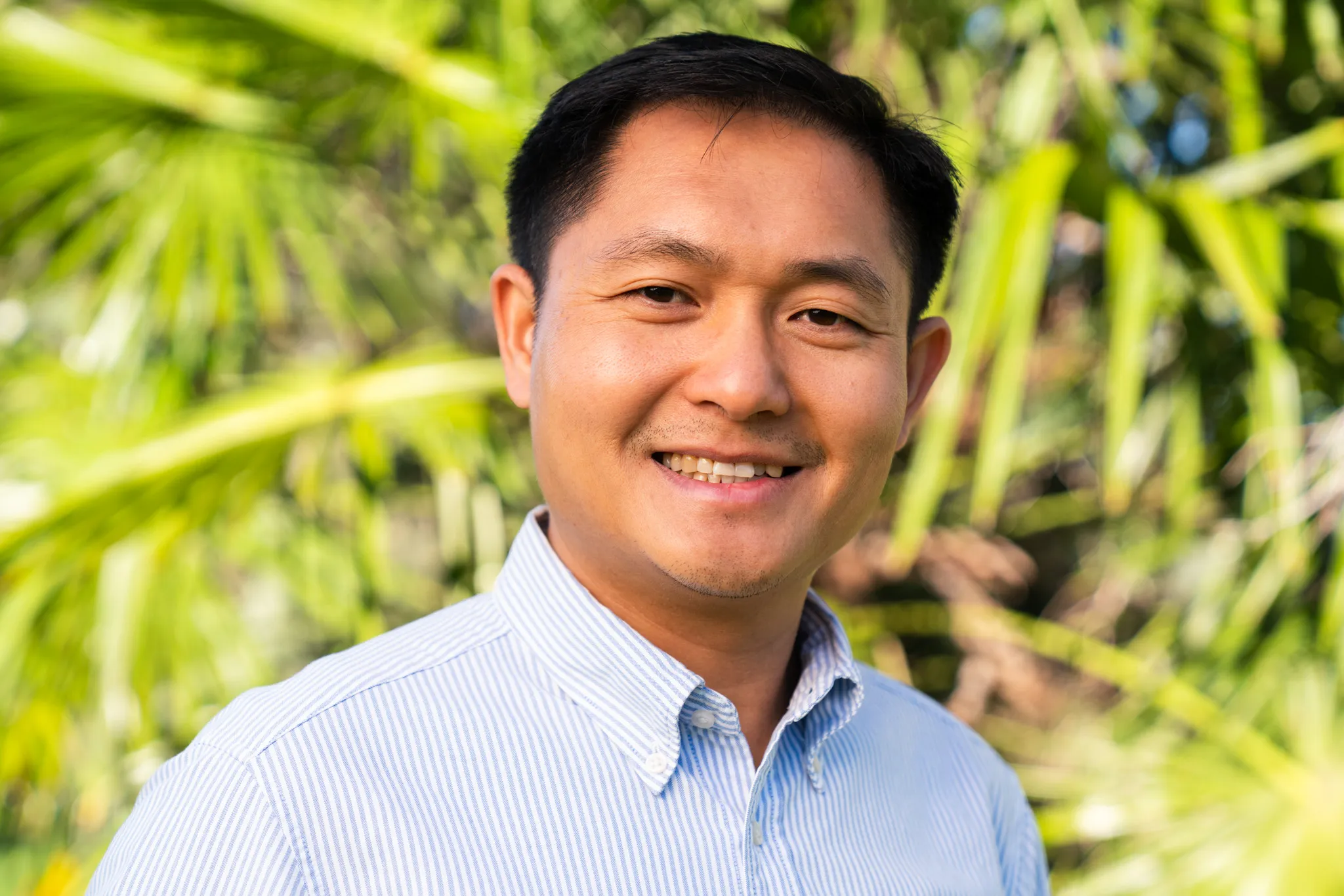 A man wearing a business shirt stands smiling with a green palm bush in the background.