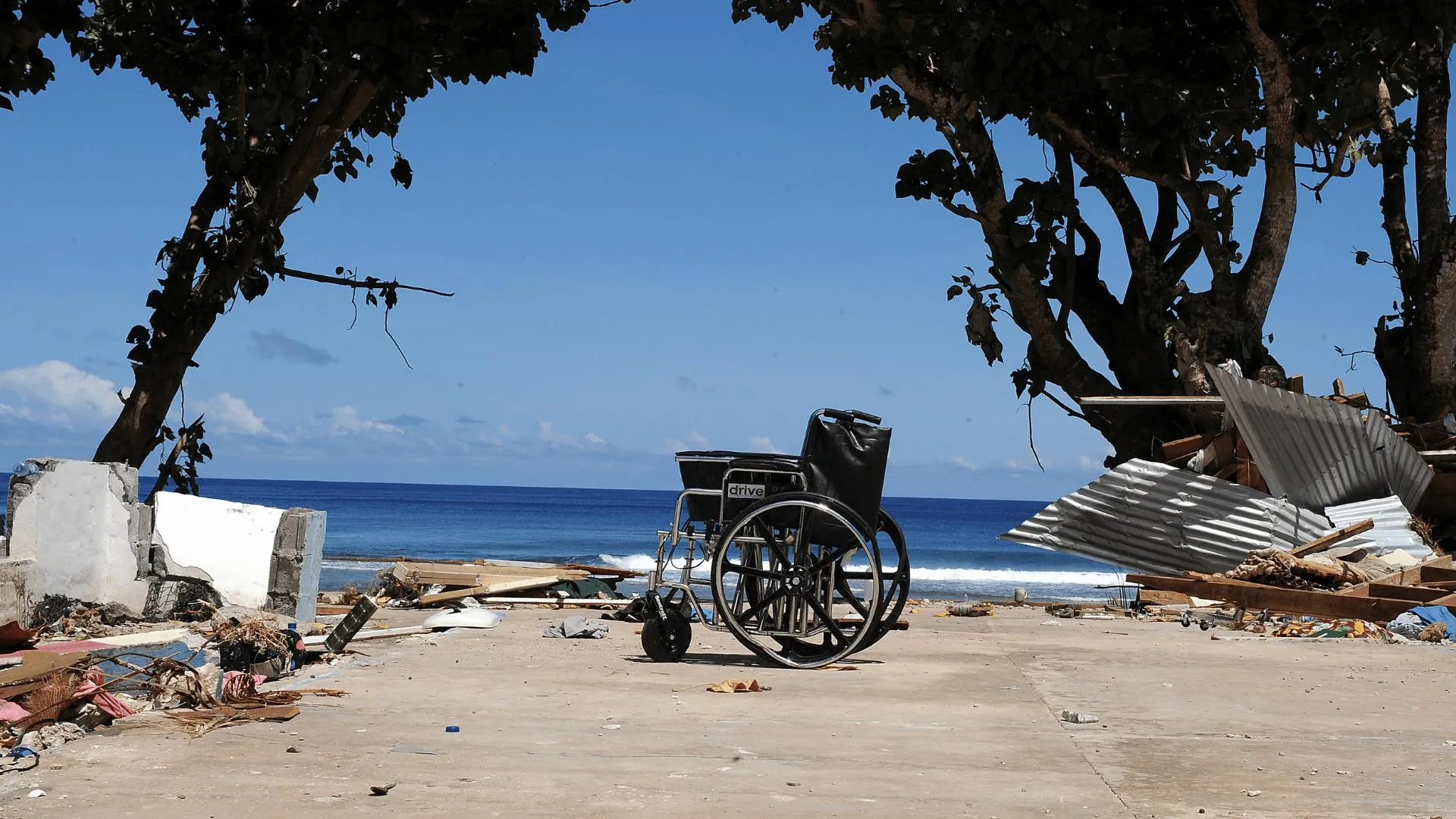 An empty wheelchair sits amid storm debris and collapsed structures on a coastal area, with the ocean in the background - highlighting the vulnerability of people with disabilities during climate disasters.