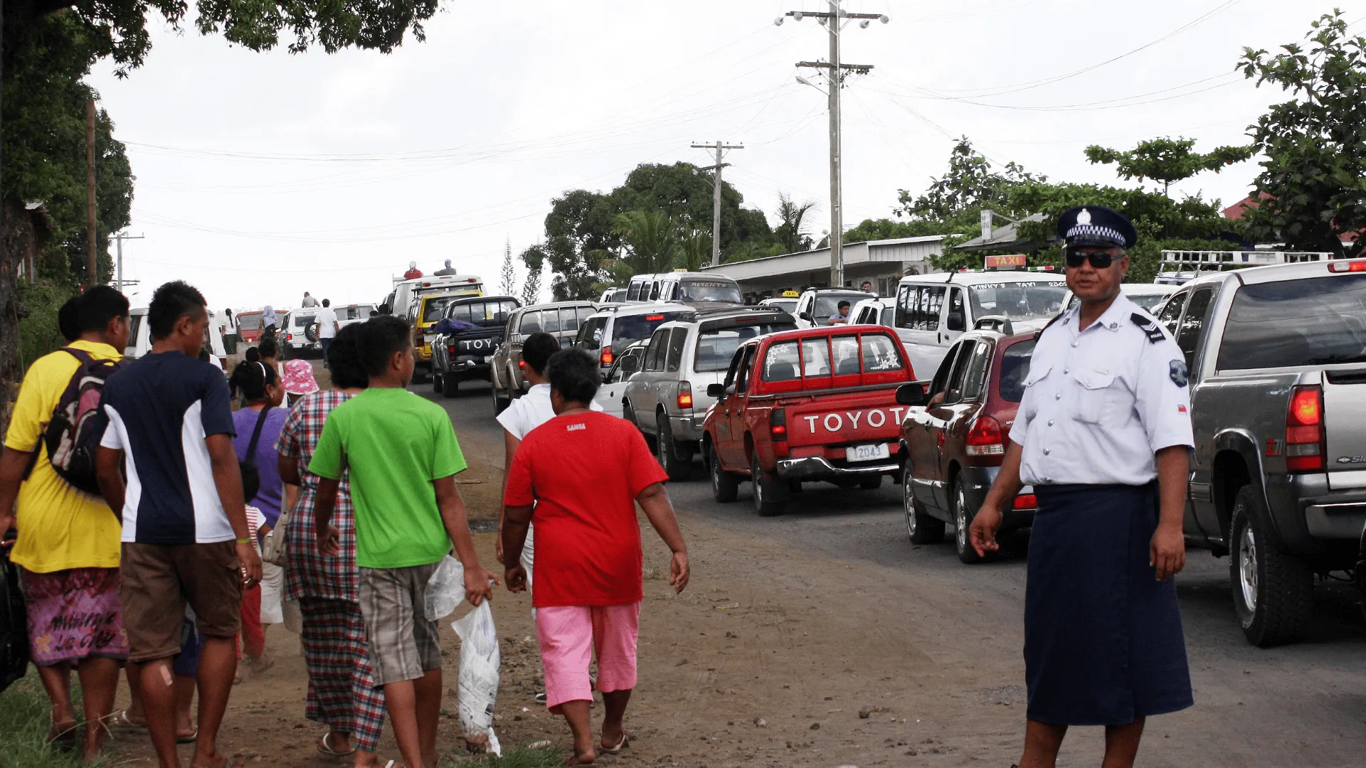 A crowd of people, including families and children, walk along a congested roadside as vehicles line up during an evacuation, while a uniformed police officer directs traffic.