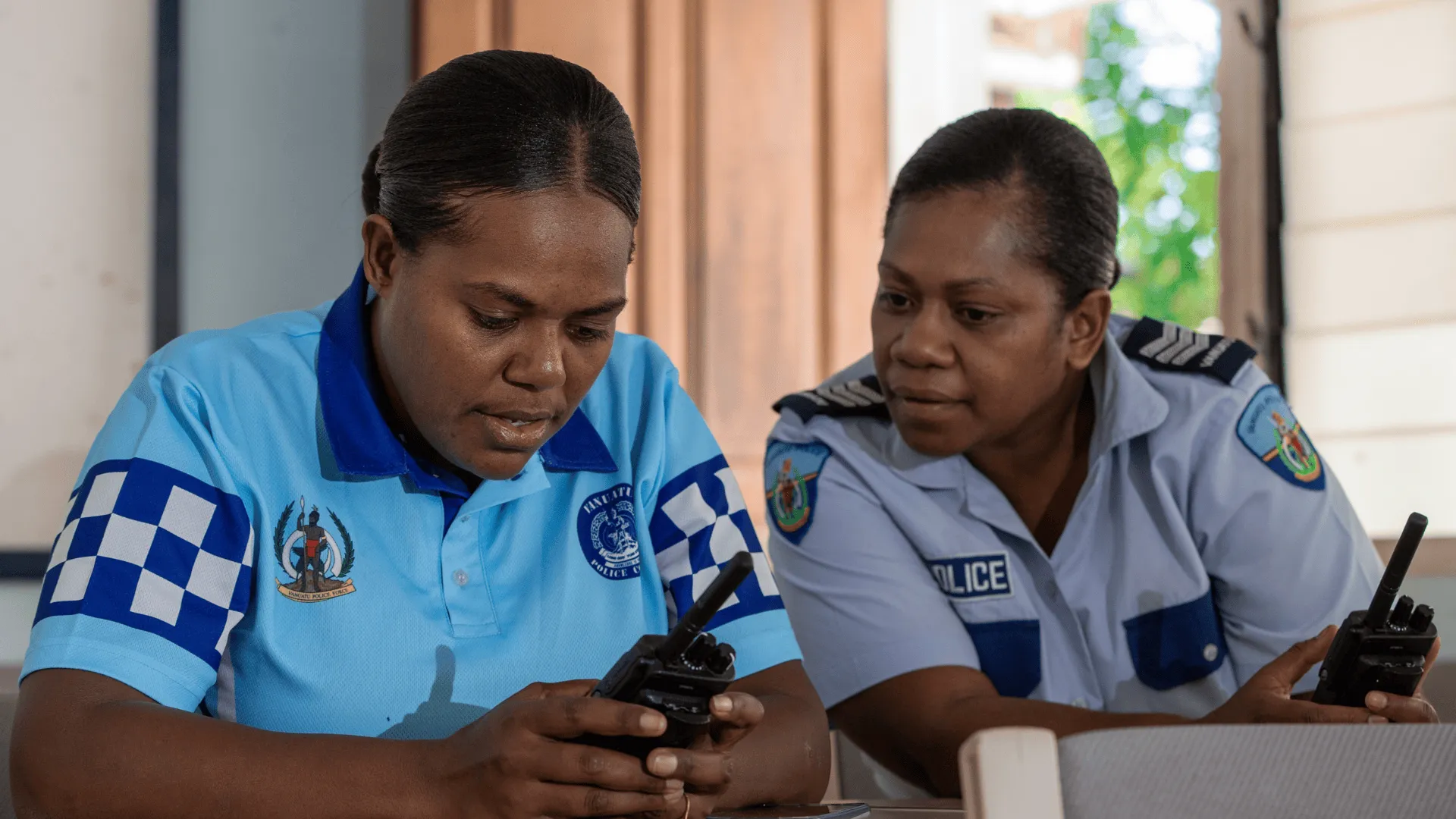 wo women in police uniforms sit together indoors, focusing on a radio communication device during a training session. One woman demonstrates while the other observes attentively.