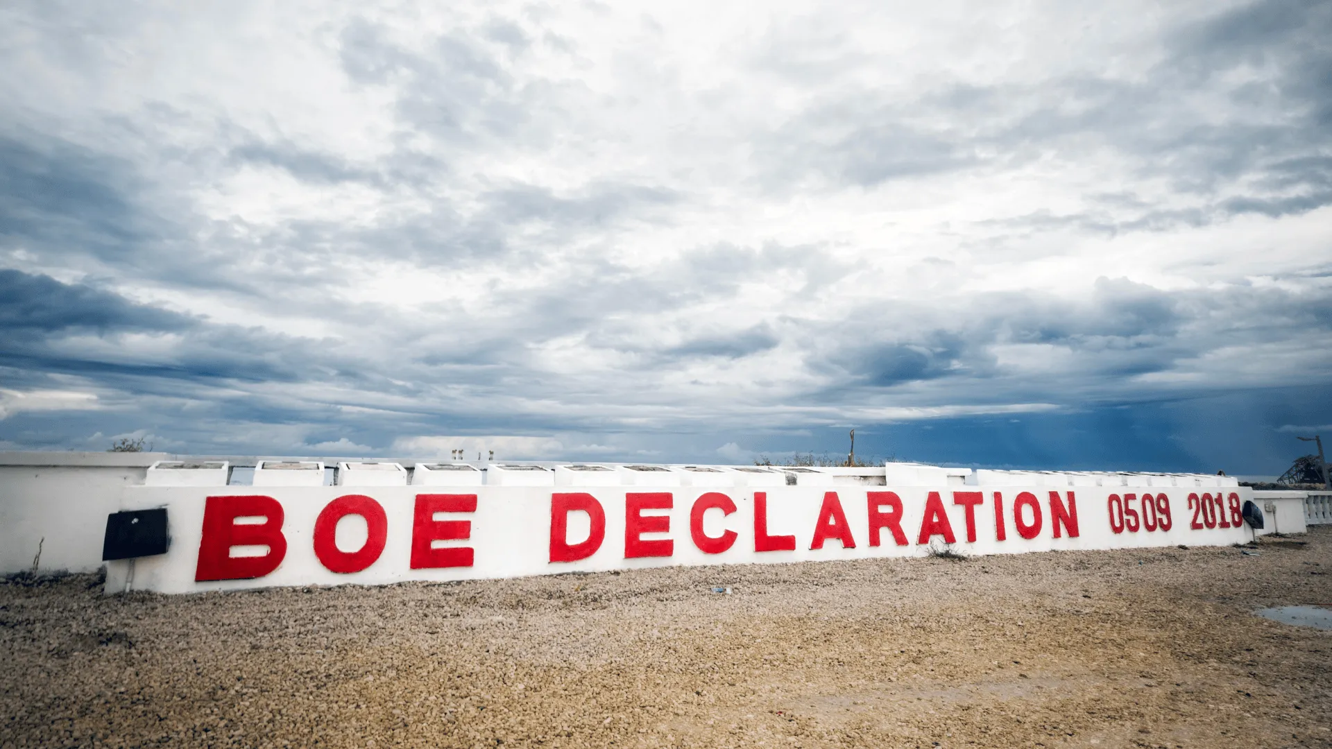 A long white seawall with the words “BOE DECLARATION 0509 2018” painted in large red letters, set against a dramatic cloudy sky and a rocky shoreline.