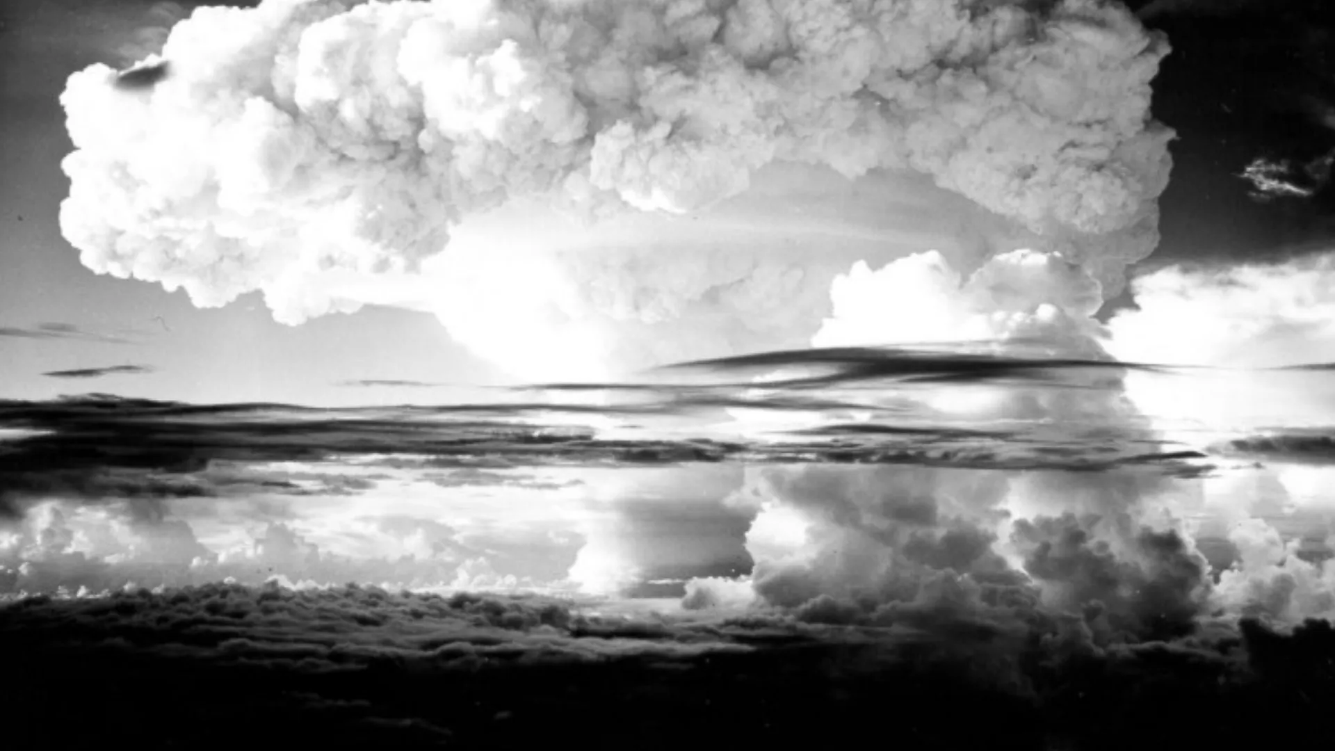 A black-and-white photograph showing a massive mushroom cloud rising above layered clouds during a nuclear test explosion.