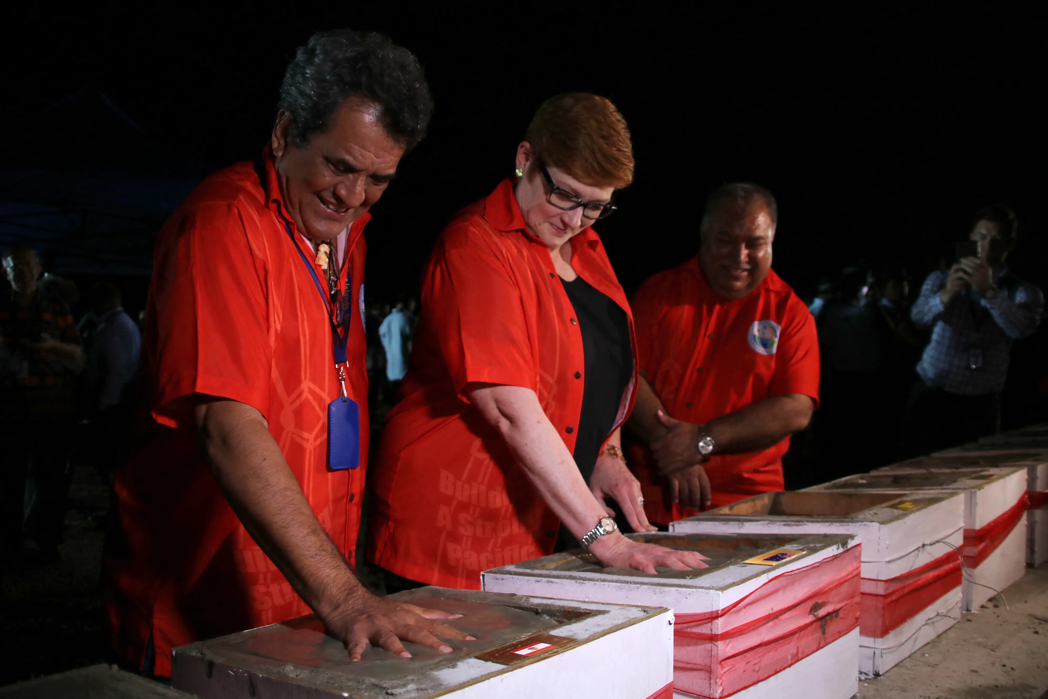 Three adults wearing matching red shirts place their hands on rectangular concrete blocks during a ceremonial event at night, with other attendees and photographers visible in the background.