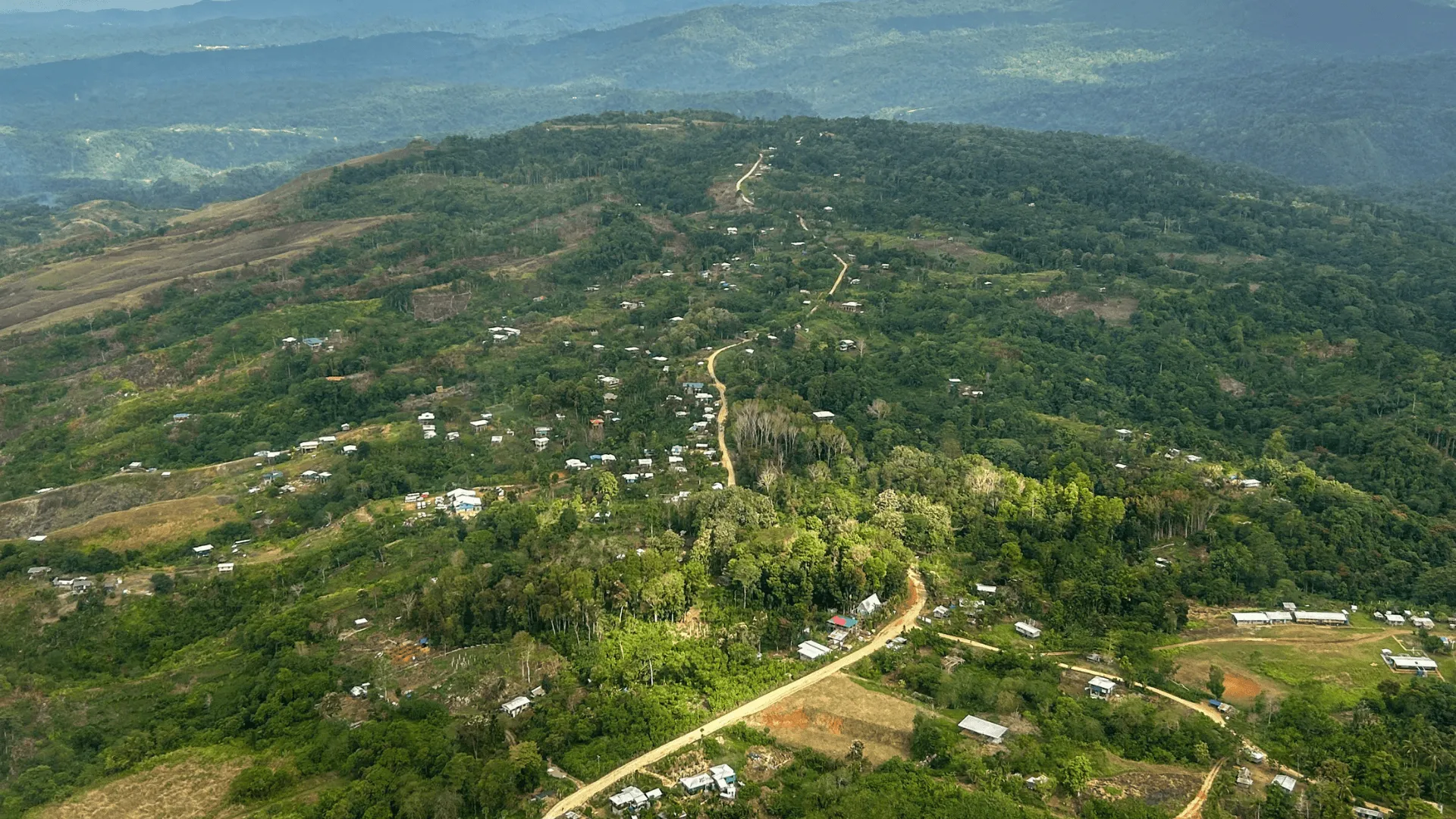 Aerial view of a rural Pacific Island village scattered along a winding road through forested hills and cleared farmland.