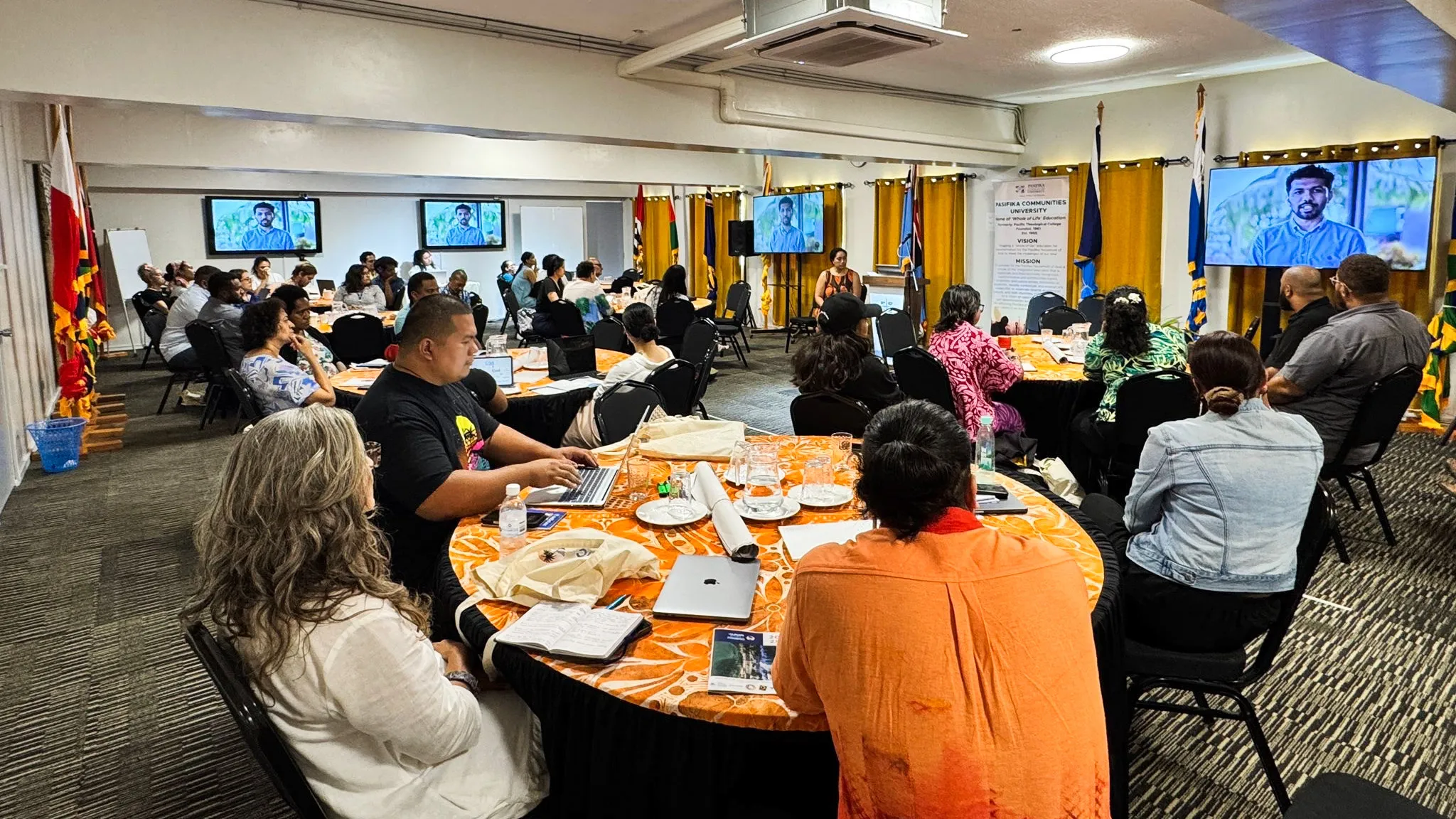 A group of people sit around tables watching a man presenting on a television screen.