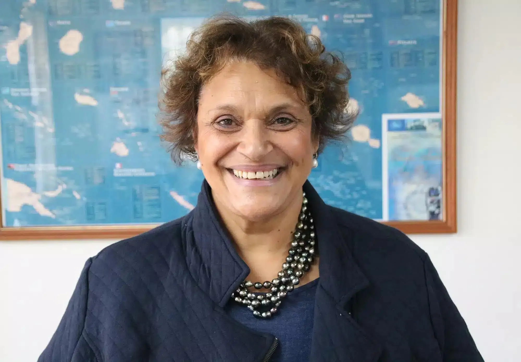 Smiling woman with short curly hair wearing a dark jacket and a layered pearl necklace, standing in front of a framed world map on the wall.