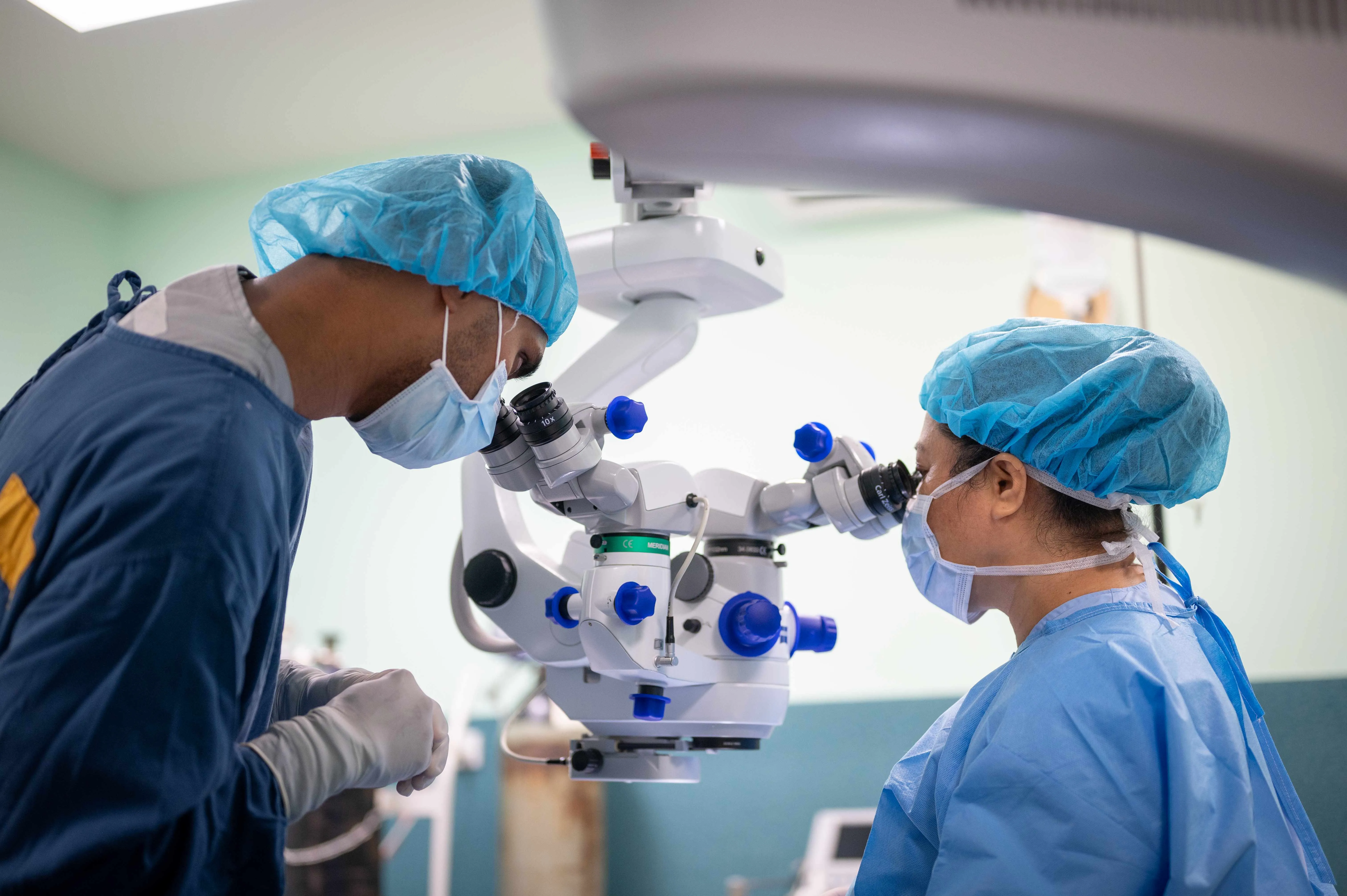 Two surgeons wearing blue surgical caps and masks use a microscope during a medical procedure in an operating room.
