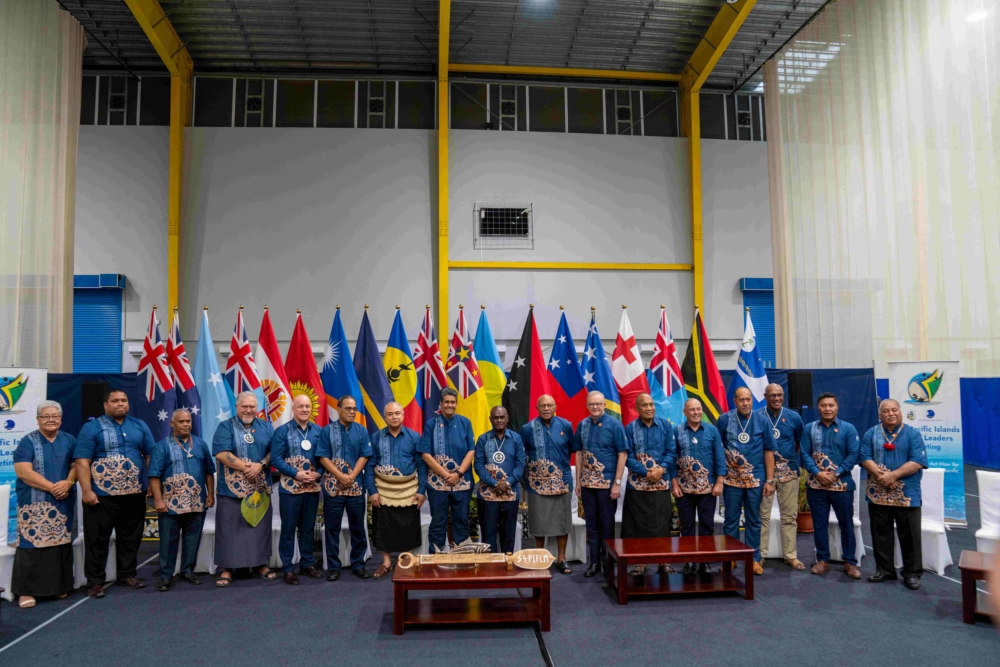 A row of Pacific Islands Forum member flags stands behind standing delegates wearing blue shirts at a formal regional meeting indoors.