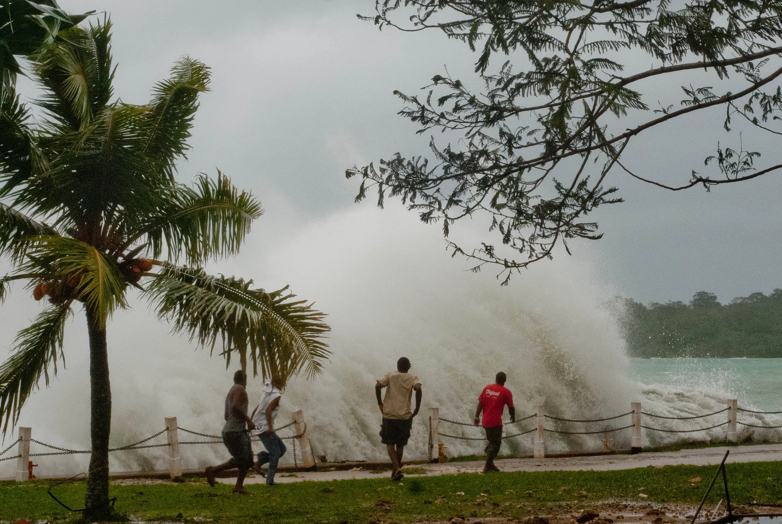 People run along a waterfront as large storm waves crash over a seawall, with palm trees bending in strong winds during a cyclone.