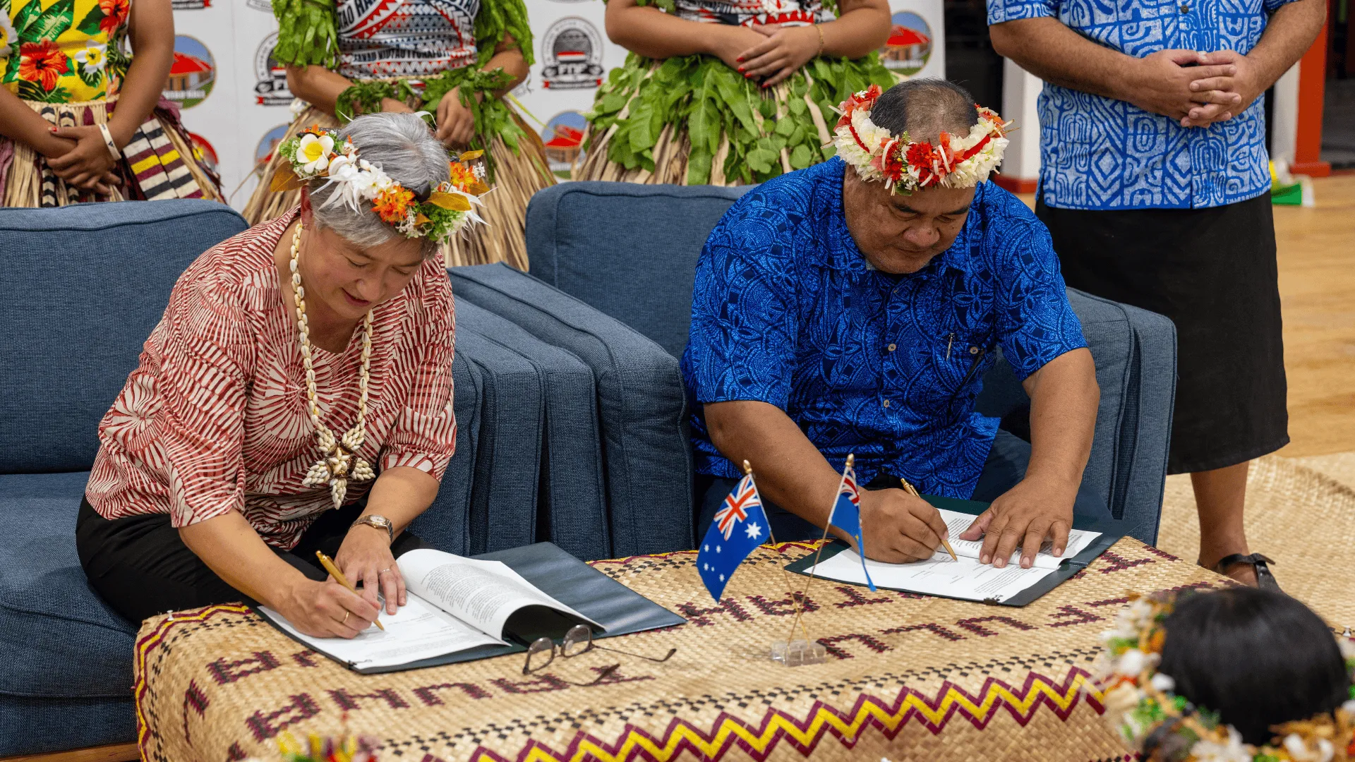 Two officials wearing floral garlands sign documents at a woven mat-covered table, with small Australian and Tuvalu desk flags in the foreground and people standing behind them