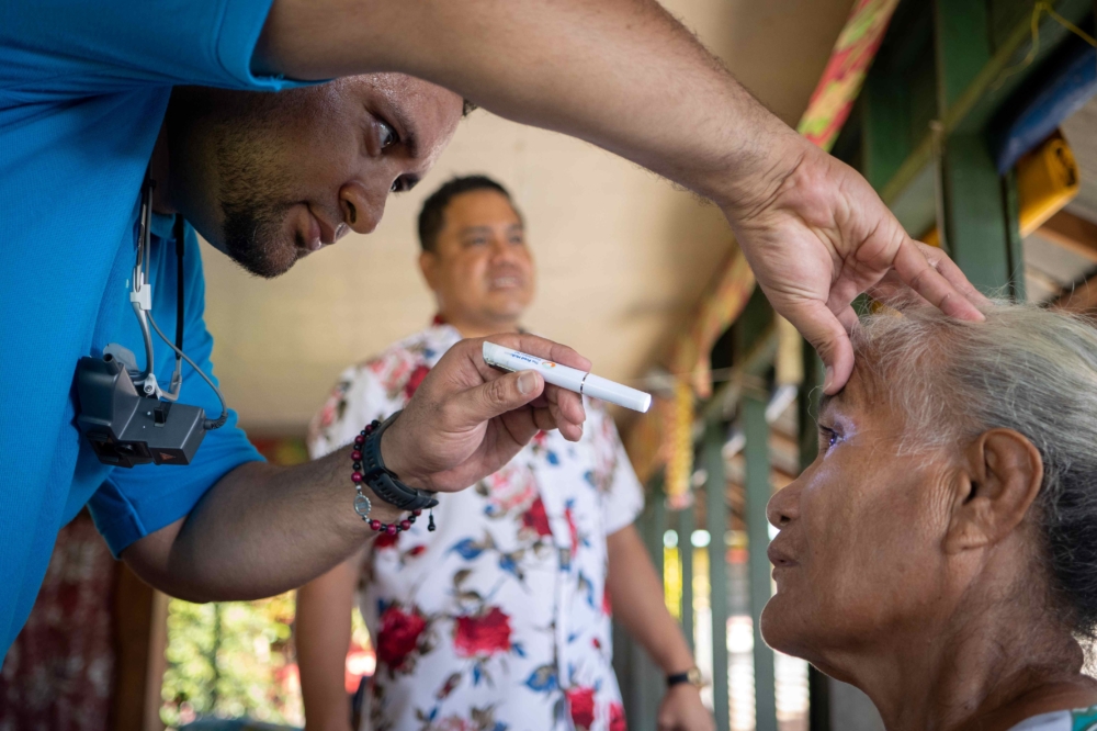 A health worker examines an elderly woman’s eye using a penlight during a community eye health check, with another staff member observing in the background.