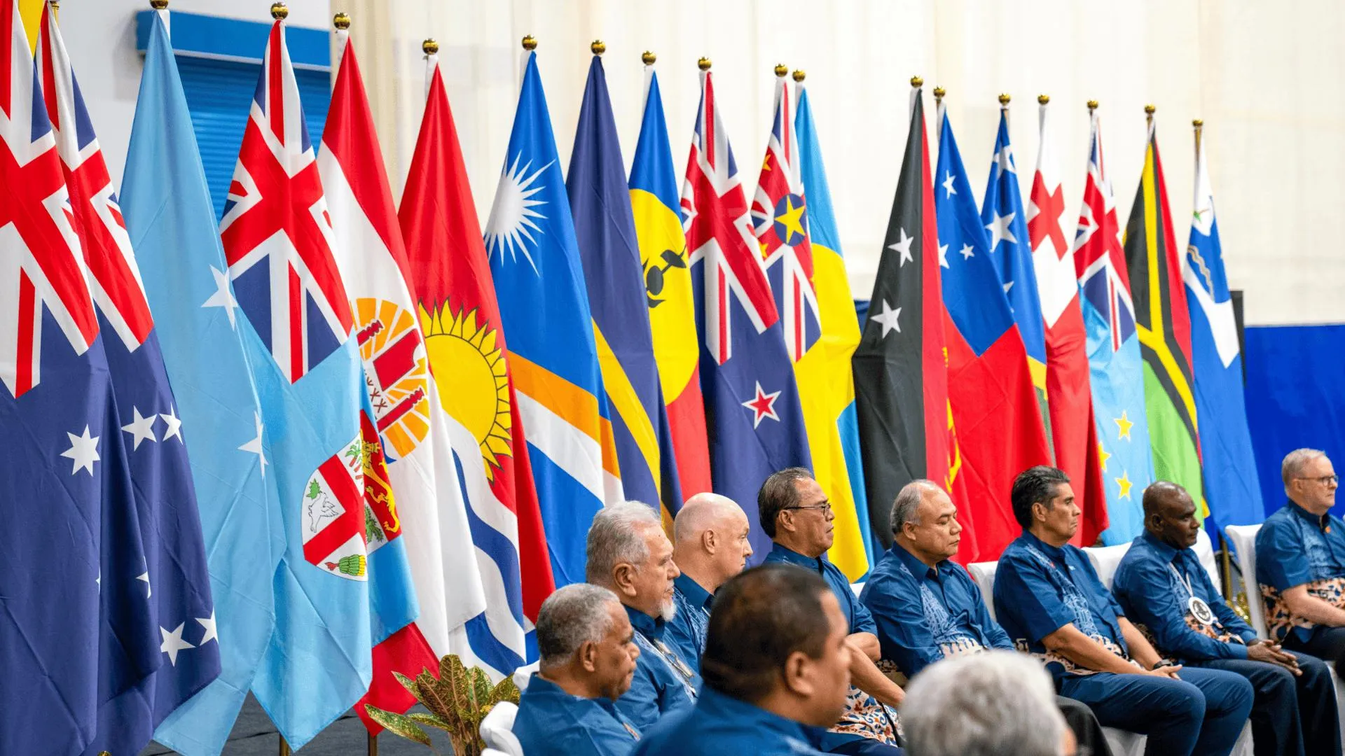 Pacific leaders sit in a row at a formal meeting, with national flags of Pacific Island countries displayed behind them.