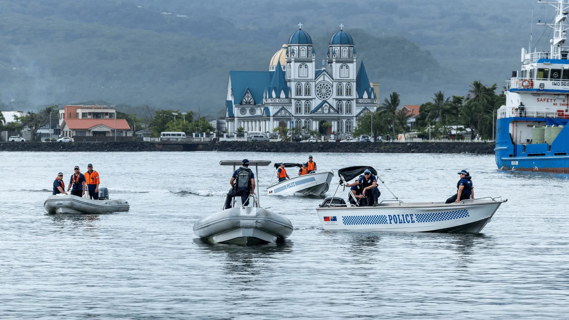 Several police boats patrol a harbour, with officers wearing life jackets, as a large church and coastal town sit against a mountainous backdrop.