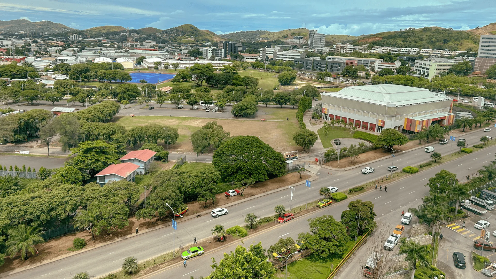 Aerial view of a leafy urban area featuring a large modern building beside a multi-lane road with light traffic, surrounded by trees, low-rise buildings, blue sports courts, and rolling hills in the background.