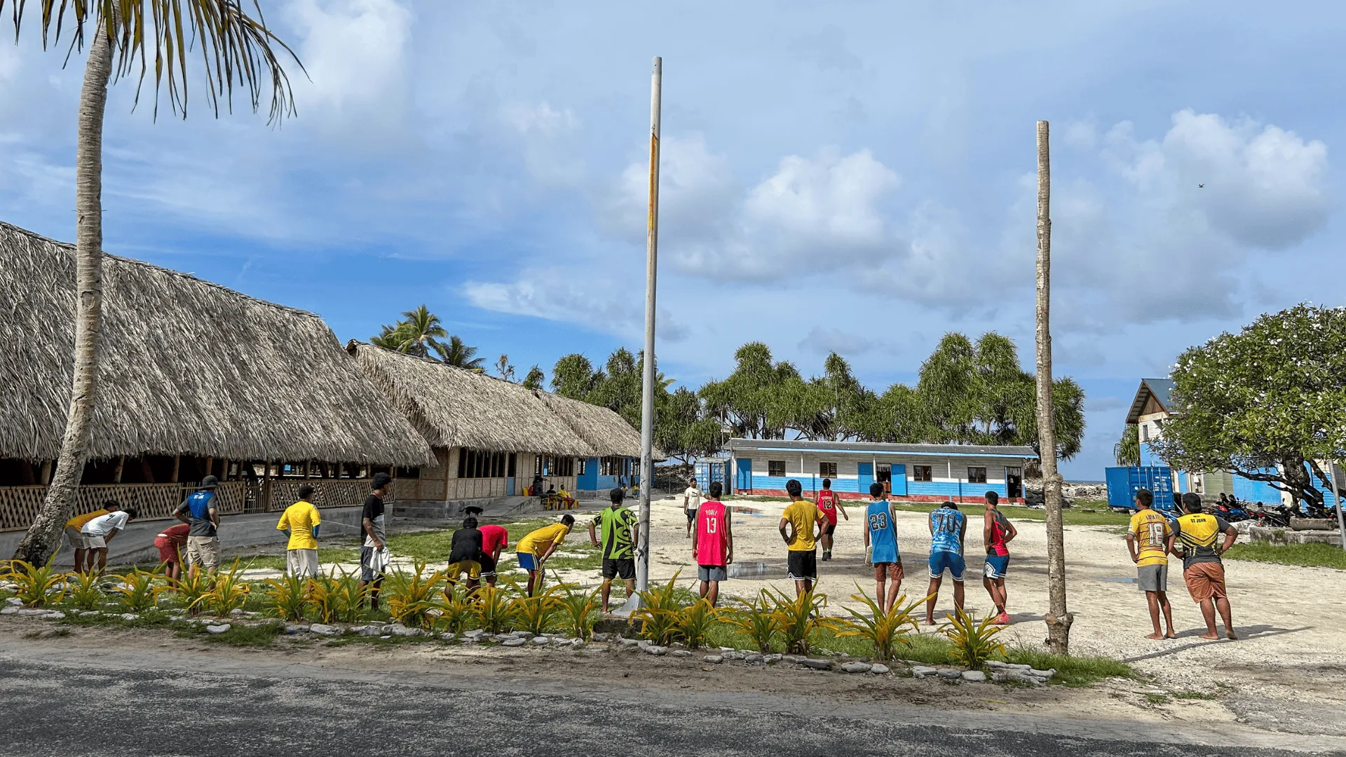 Young men stand and watch a volleyball game on a sandy court in a Pacific island village, with thatched buildings and palm trees nearby.