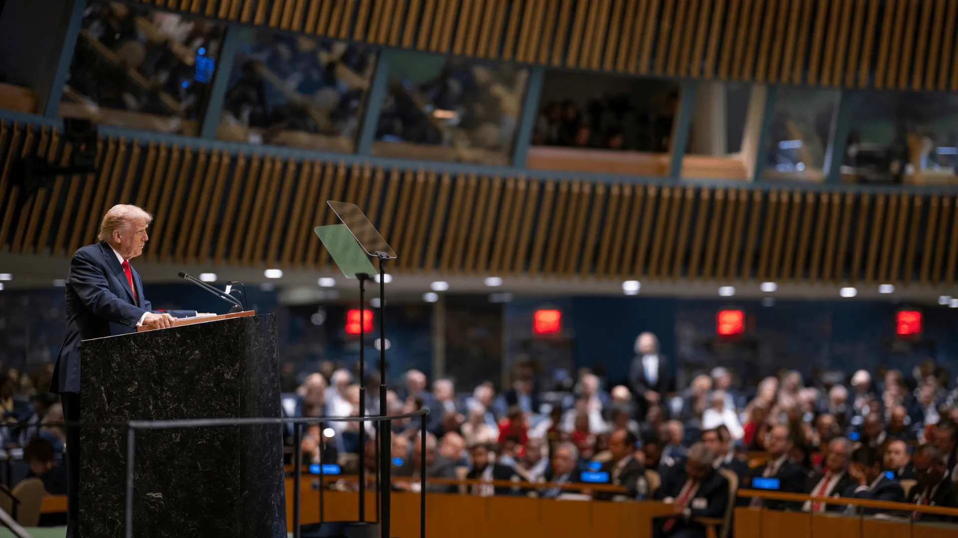A suited speaker stands at a large podium addressing an assembly inside a grand international chamber, with audience members seated in the background.