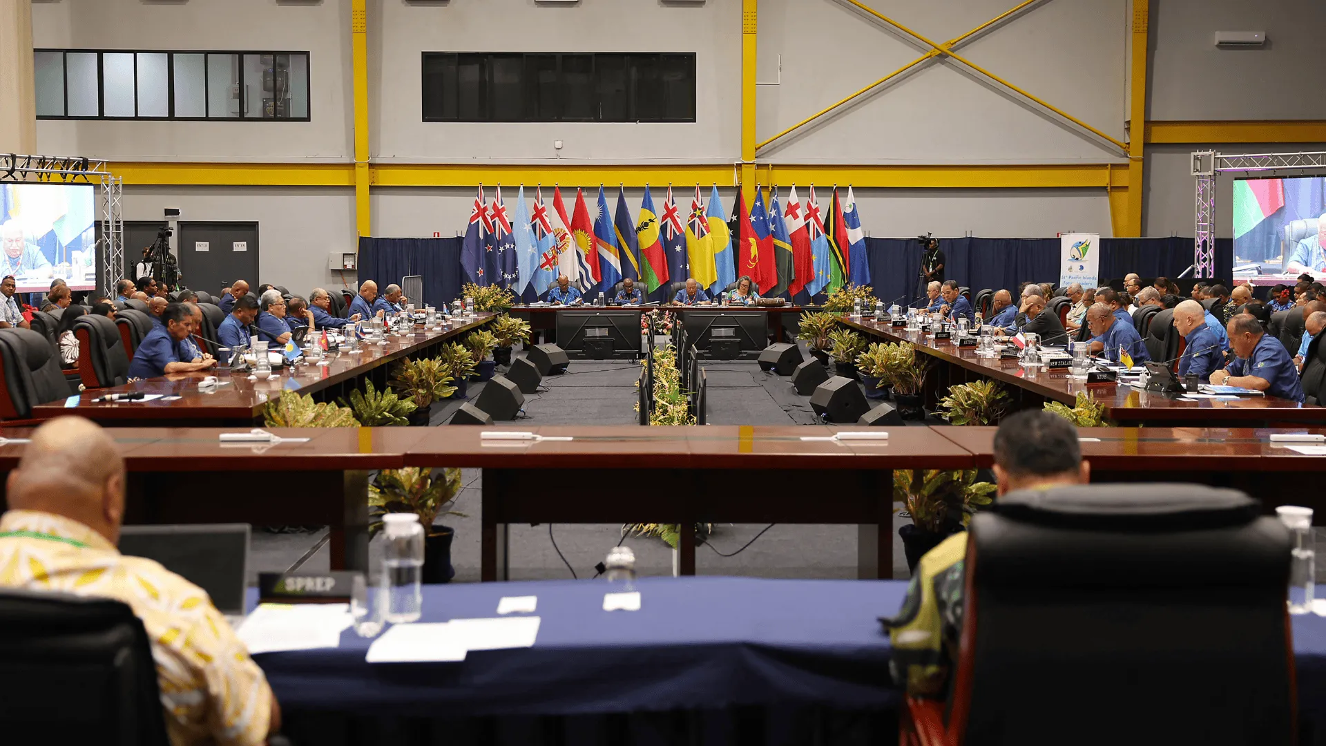 Delegates sit around long conference tables during a regional meeting, with Pacific Island nation flags displayed behind a central panel of speakers at the front of the hall. Large screens show the proceedings as officials listen and take notes in a large indoor conference venue.