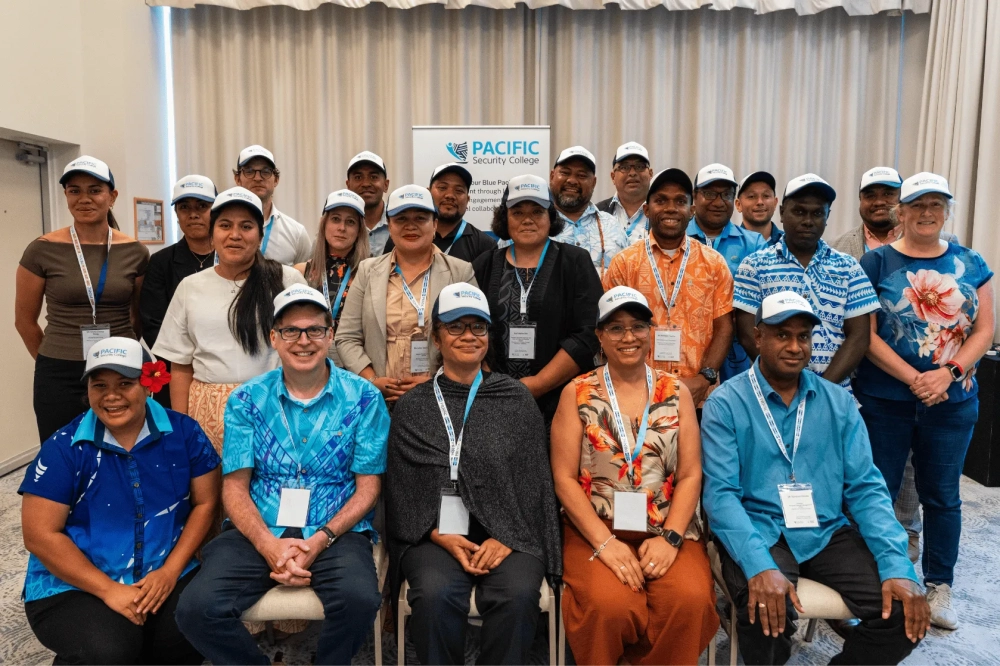 A group of around 18–20 participants pose for a group photo indoors at a workshop, seated and standing in rows. They wear matching light blue caps and conference lanyards, with a banner for the Pacific Security College visible behind them.