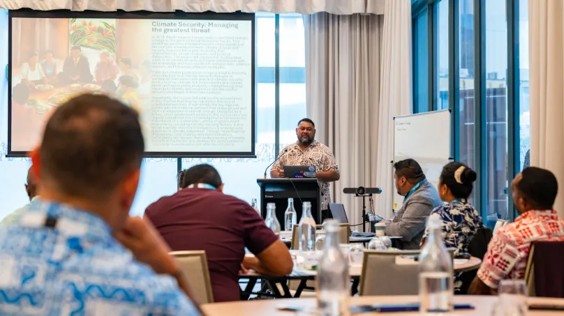 A man stands at a lectern delivering a presentation to a seated audience in a conference room. A slide titled “Climate Security: Managing the greatest threat” is projected on a screen behind him, while attendees sit at tables with water bottles and notebooks, listening attentively.