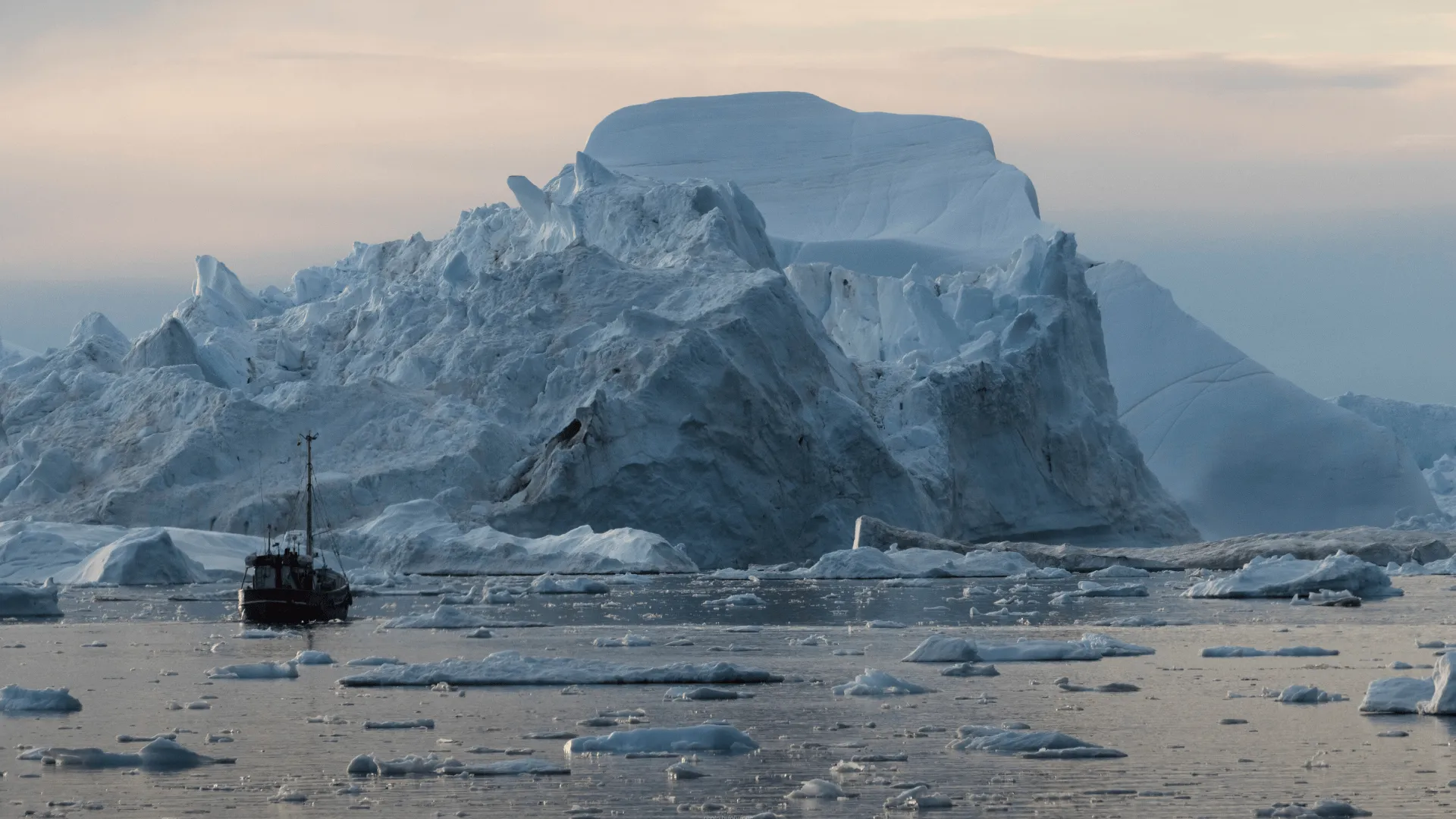 A small boat navigates icy waters beside a massive iceberg surrounded by floating ice fragments under a soft, cloudy sky.