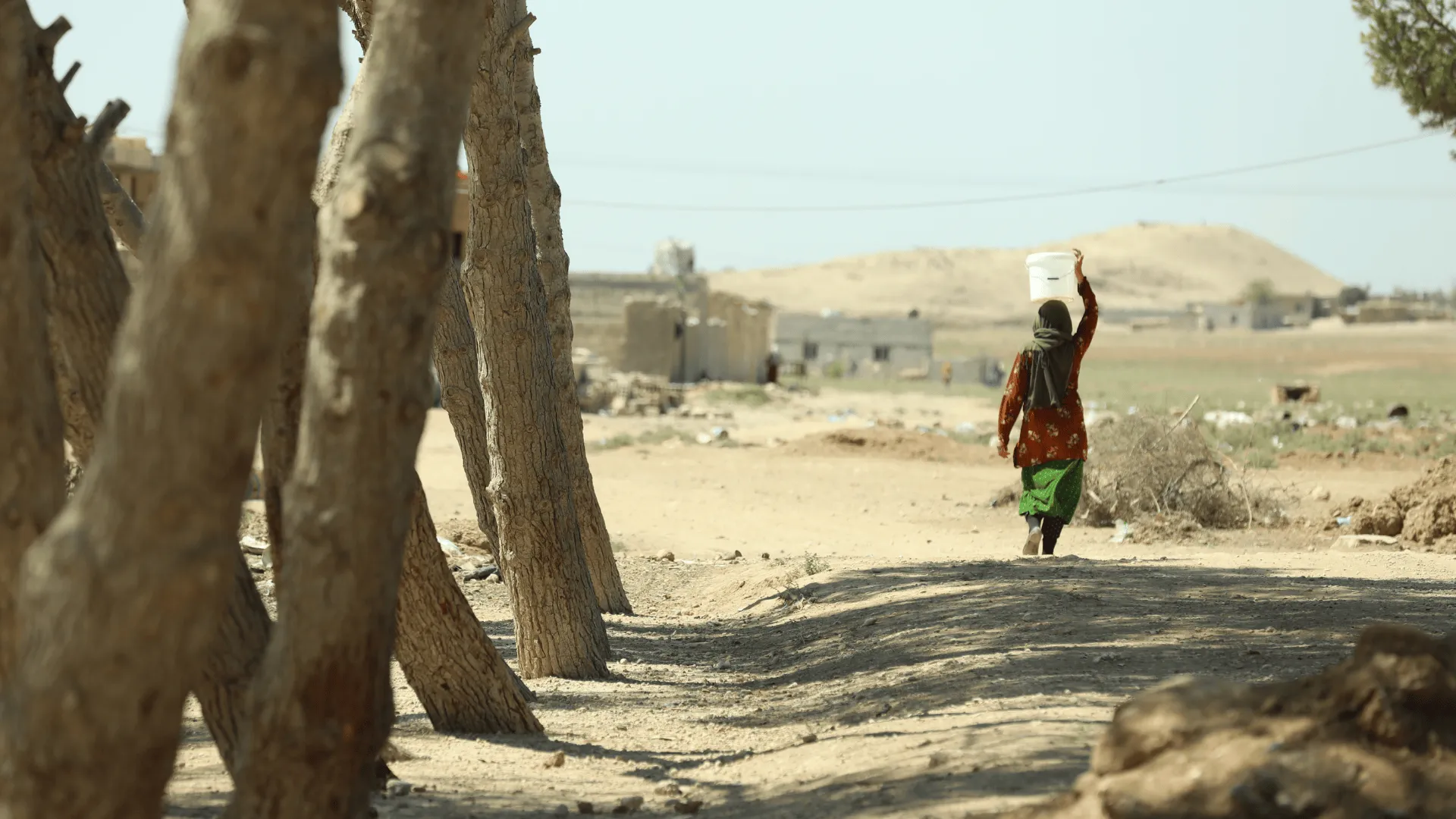 A woman walks along a dusty rural path carrying a white water container on her head, with sparse trees, low buildings, and dry hills in the background.