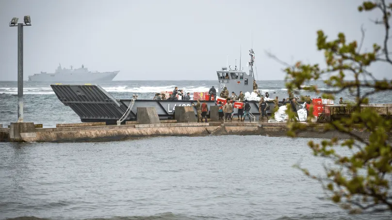 A small military landing craft is docked at a concrete pier as uniformed personnel unload boxes and supplies, some carrying red containers; the sea is rough, and a larger naval ship is visible in the hazy distance offshore.