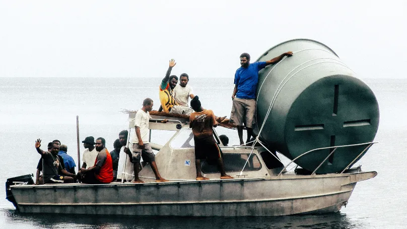 A group of men crowded onto a small motorboat at sea, transporting a large cylindrical water tank secured on the deck; one man stands on top of the tank while others sit and hold onto the boat.