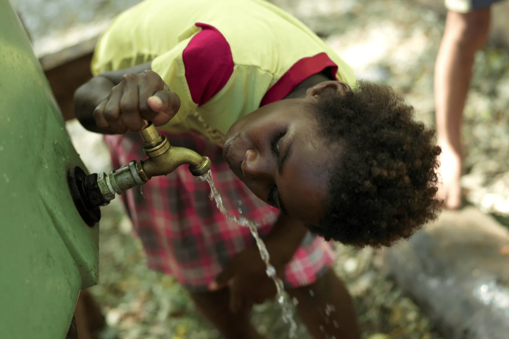 A young child bends down to drink water directly from a running outdoor tap attached to a green tank, with water splashing as they cup their mouth close to the stream.
