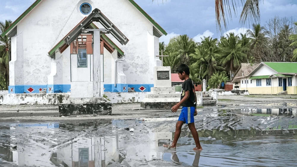 A young boy walks barefoot through shallow floodwater in front of a small white church in a tropical village, with palm trees and houses in the background reflected in the water.
