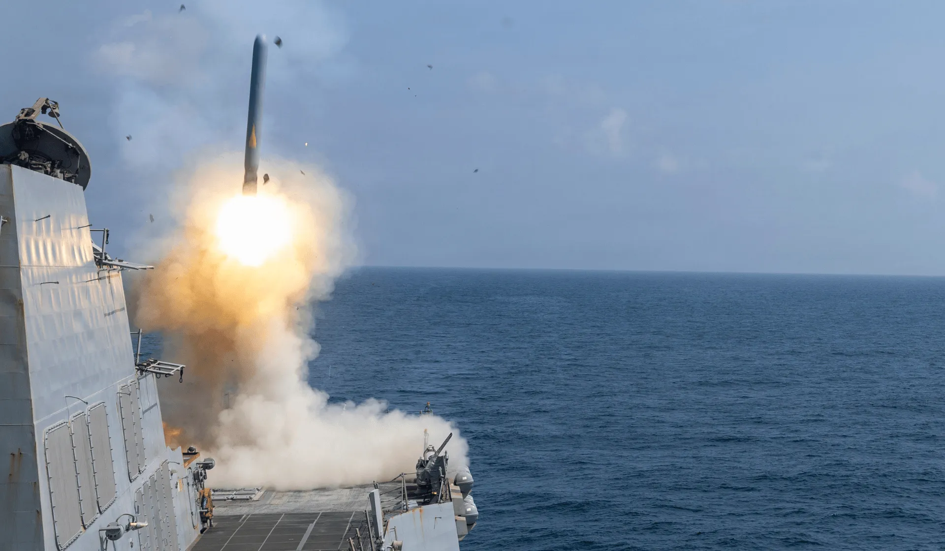 A missile launches vertically from a naval warship at sea, with a bright burst of flame and thick smoke erupting from the launch system on the ship’s deck. The vessel is surrounded by open ocean under a cloudy sky as debris and smoke trail behind the ascending missile.