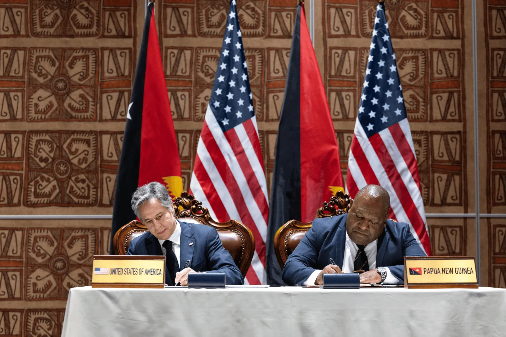 Two officials sit side by side at a table signing documents, with nameplates reading “United States of America” and “Papua New Guinea.” Behind them are alternating national flags of both countries and a patterned wall backdrop, suggesting a formal bilateral agreement ceremony.