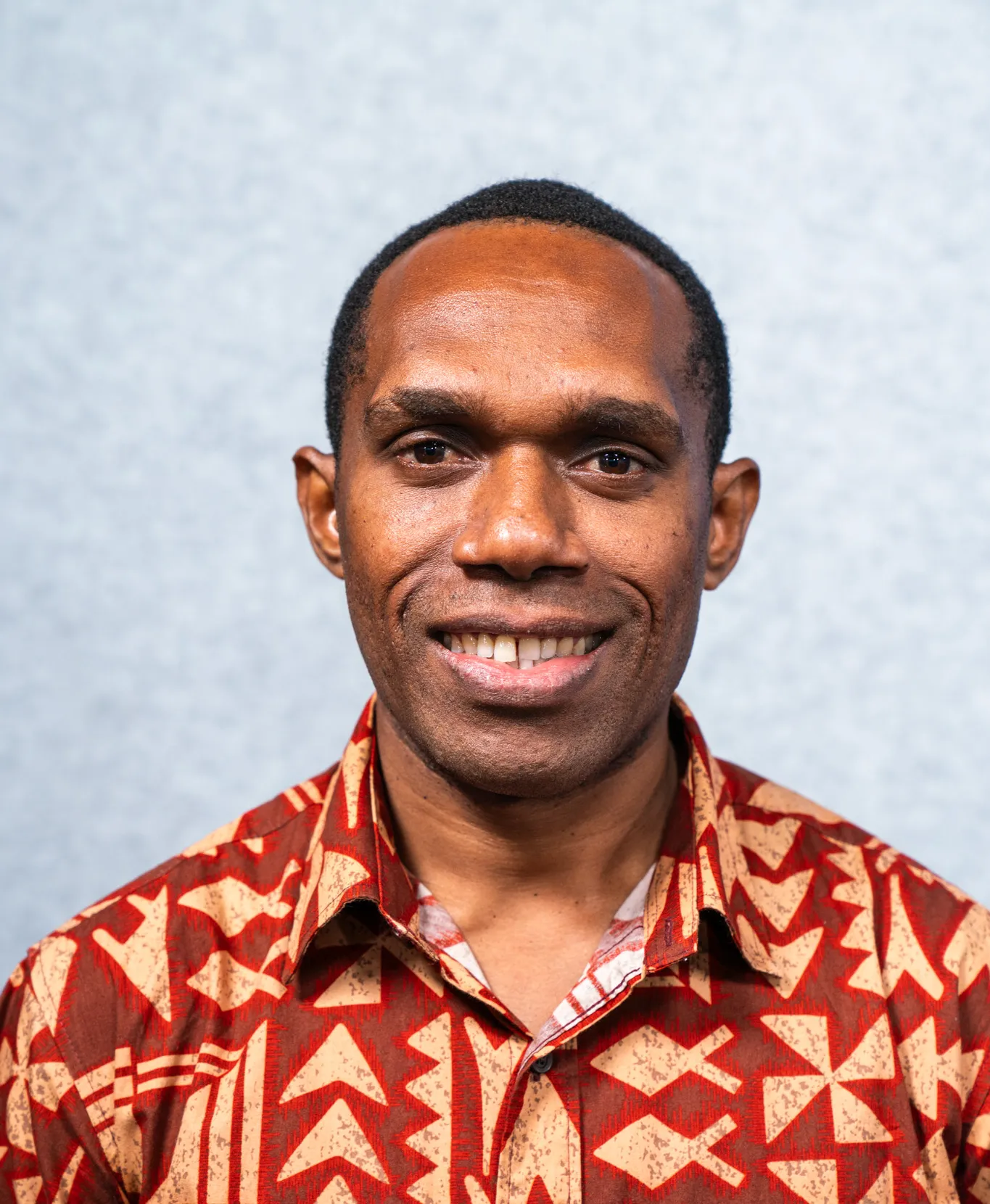 A man stands smiling wearing a red shirt with yellow shapes on it.