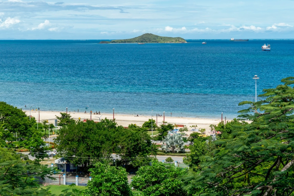 Wide coastal view of a sandy beach with people walking and swimming, bordered by lush green trees and a seaside road, with a small island offshore and several ships anchored in the calm blue ocean under a partly cloudy sky.