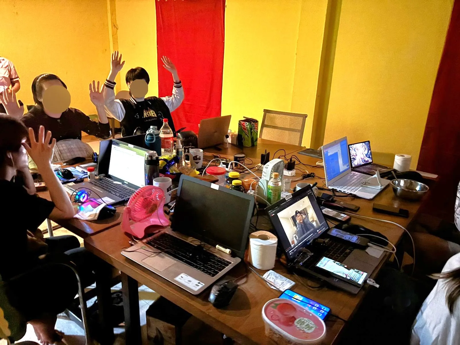 Group of people seated around a large table covered with laptops, phones, drinks, and cables, several raising their hands with their faces blurred out in a brightly colored room.