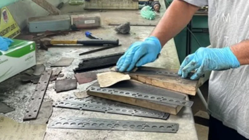 A person wearing blue gloves works at a cluttered bench, handling layered material samples and metal strips with tools scattered nearby.
