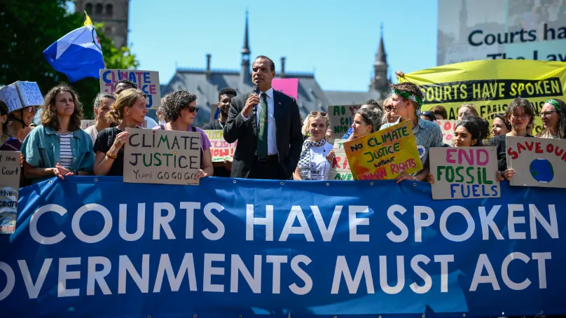 A group of climate activists gather at a rally outdoors, holding signs that read “Climate Justice,” “End Fossil Fuels,” and “No Climate Justice Without Human Rights.” At the centre, a man in a suit speaks into a microphone. A large blue banner in the foreground reads “Courts Have Spoken Governments Must Act,” while a yellow banner behind the crowd says “Courts Have Spoken.” Buildings and a clear sky are visible in the background.