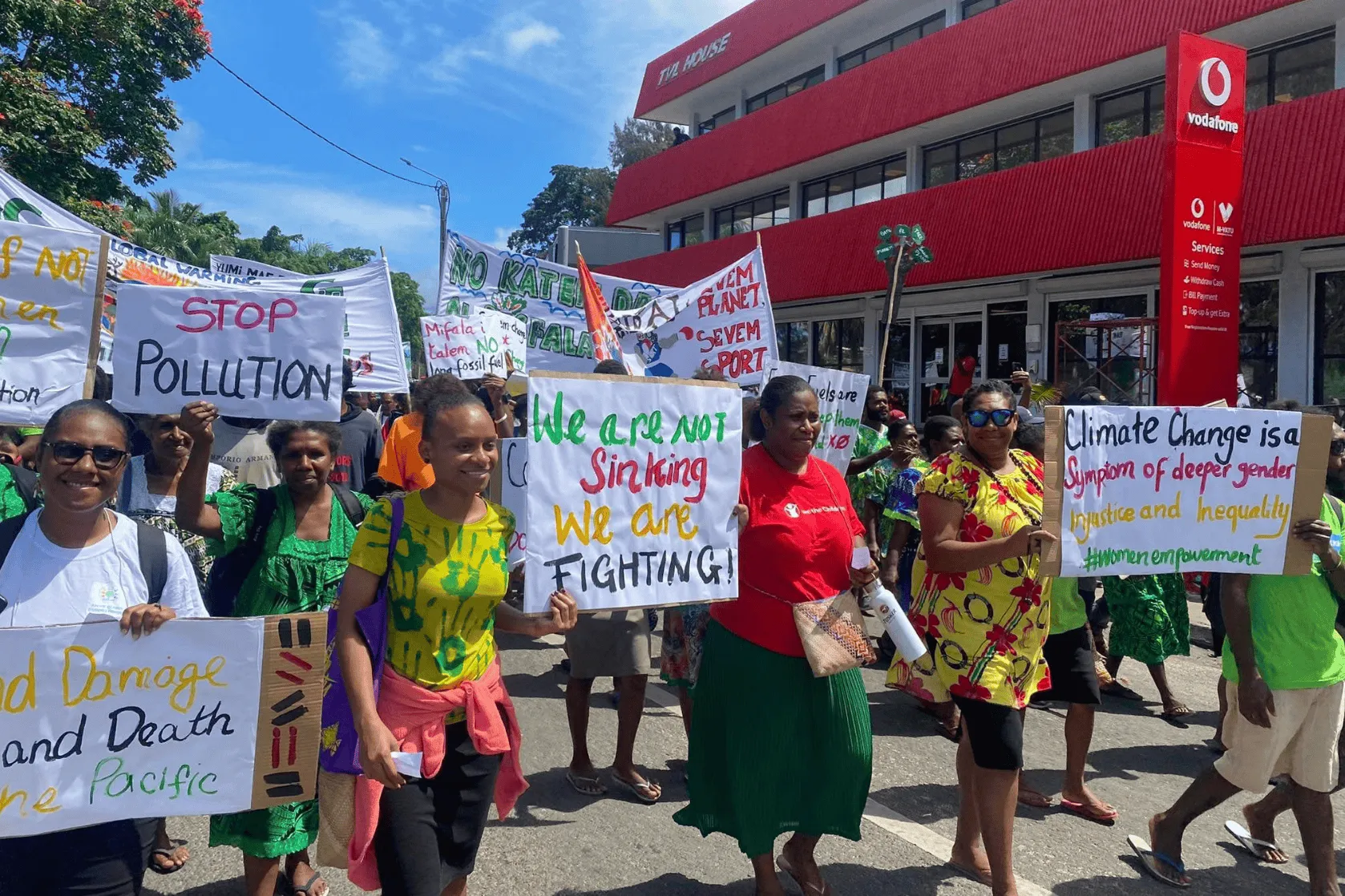 A group of people march down a street in a peaceful climate protest, holding handmade signs that read “Stop Pollution,” “We are not sinking, we are fighting,” and “Climate change is a symptom of deeper gender injustice and inequality.”