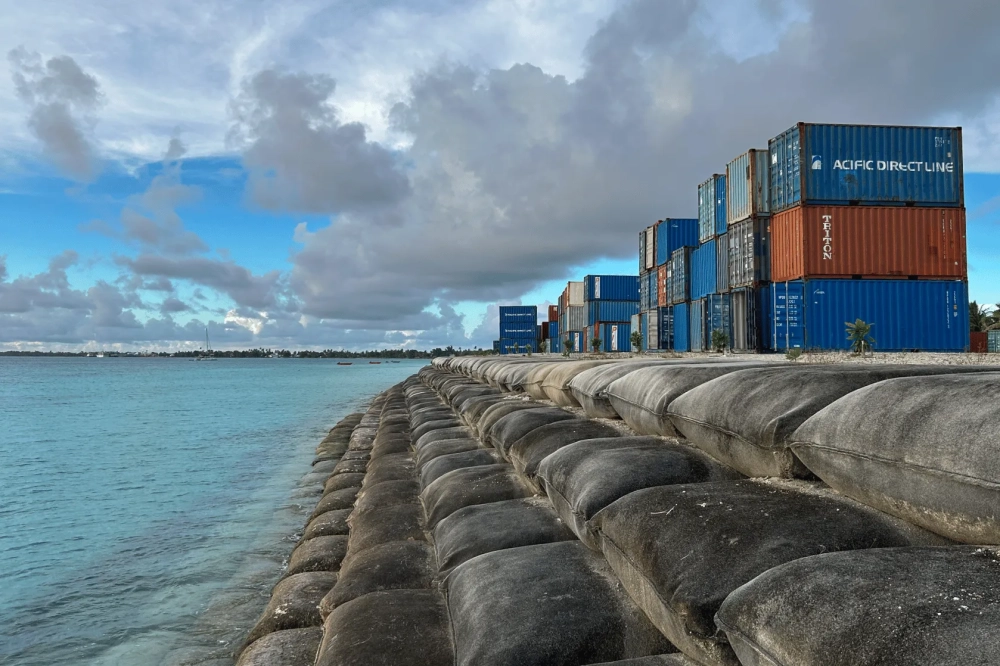 Shipping containers stacked along a coastal seawall reinforced with large sand-filled barriers, under dramatic storm clouds beside calm blue ocean water on a low-lying Pacific island.