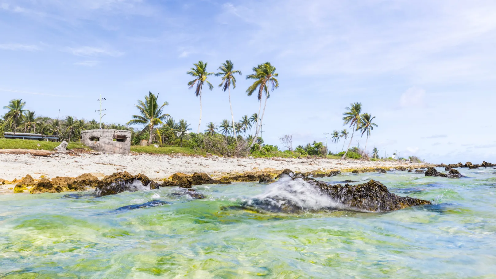 Clear shallow water washing over coral rocks along a tropical shoreline lined with palm trees, with a small concrete structure visible on the sandy coast under a bright blue sky.