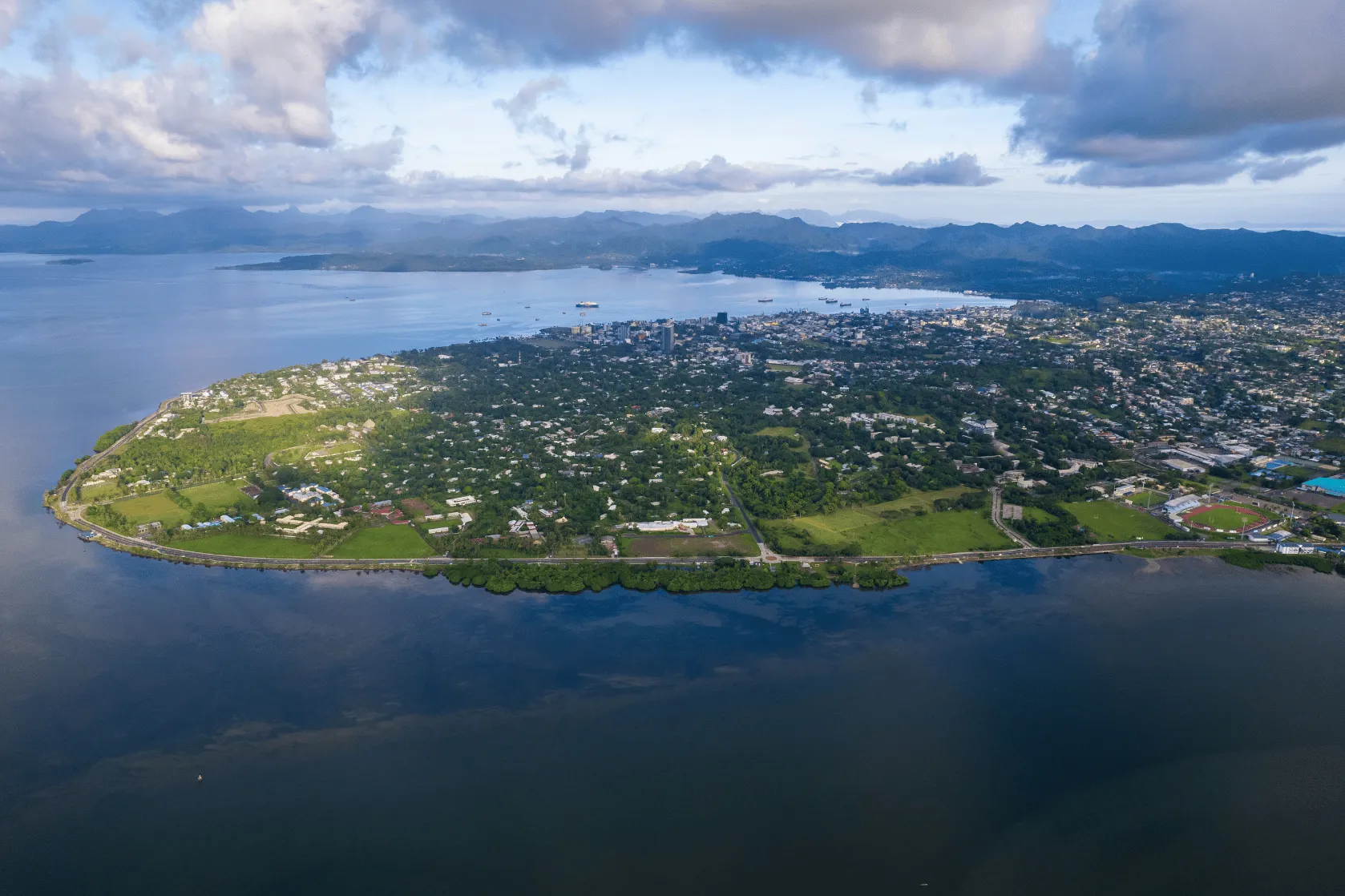 Aerial view of a coastal city surrounded by ocean and green hills under dramatic clouds, with a curved shoreline, scattered buildings, a harbour with ships, and mountain ranges stretching across the horizon.