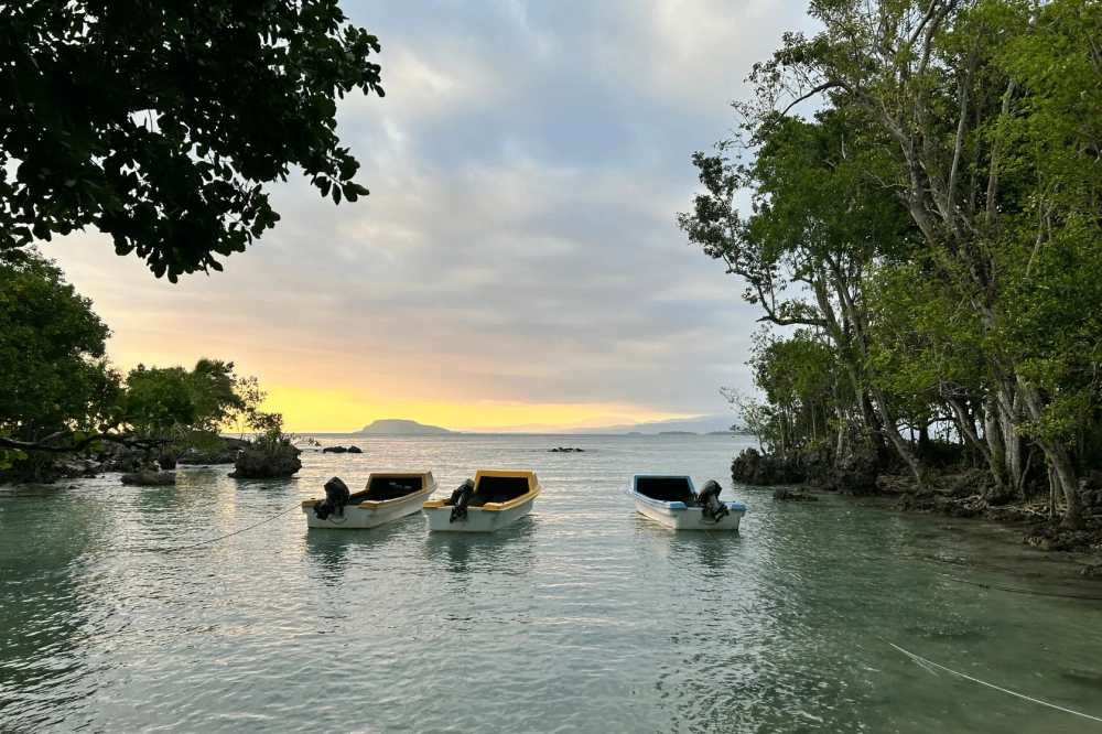 Three small motorboats float in calm, shallow water between rocky, tree-lined shores at sunset. The sky is filled with soft grey clouds with a warm yellow glow on the horizon, while distant islands are visible across the water.