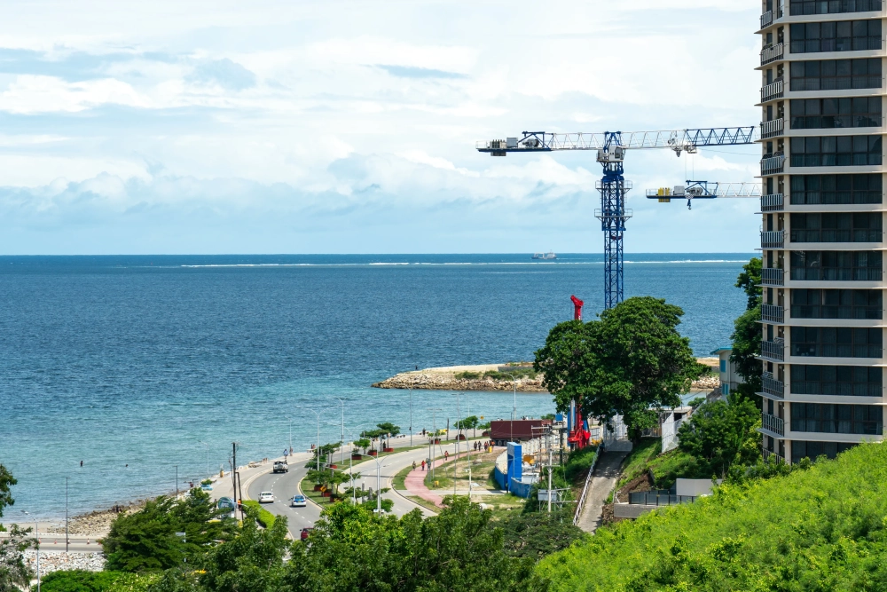 View of a coastal road curving along the shoreline beside the sea, with high-rise buildings and construction cranes overlooking the waterfront. Green vegetation covers the foreground hillside, while calm blue water stretches to the horizon beneath a cloudy sky. A small ship is visible in the distance.
