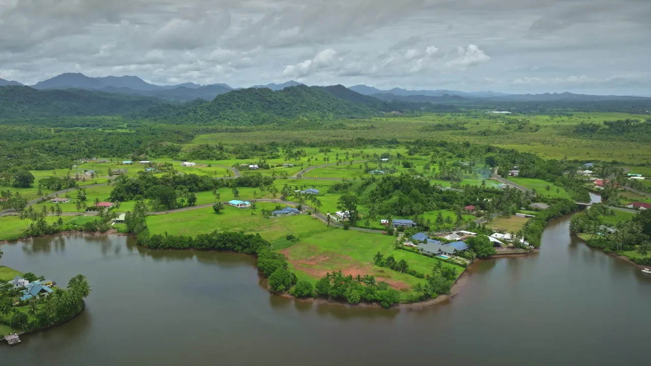 A landscape photo showing lush green fields overlooking a river with mountains in the background.