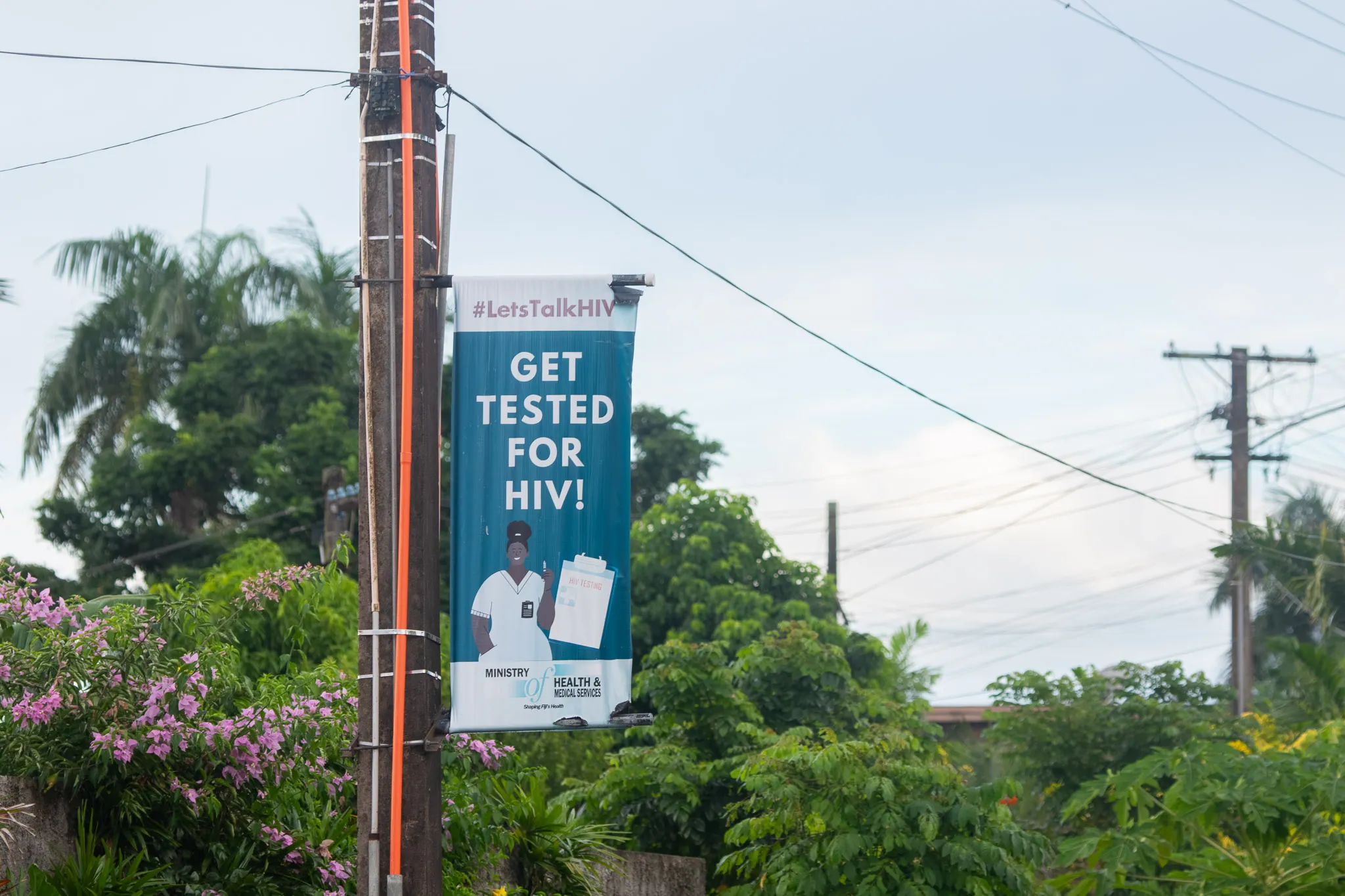 A public health banner attached to a utility pole reads “#LetsTalkHIV – Get Tested for HIV!” The sign features an illustration of a healthcare worker and HIV testing materials, with lush green vegetation, flowering plants, and power lines visible in the background under an overcast sky.