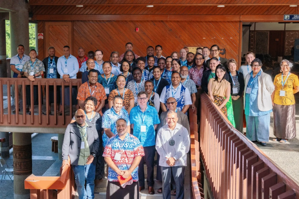 A large group of conference participants pose on a timber staircase and balcony inside a Pacific-style venue. The attendees, wearing a mix of business attire and Pacific Island clothing, stand in several rows and face the camera