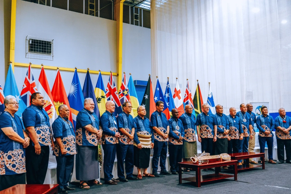 A group of Pacific leaders and officials stand side by side for a formal photograph at an indoor event venue. They are dressed in matching blue shirts featuring Pacific-inspired patterns and motifs. Behind them, a row of national flags from across the Pacific region forms a colourful backdrop.