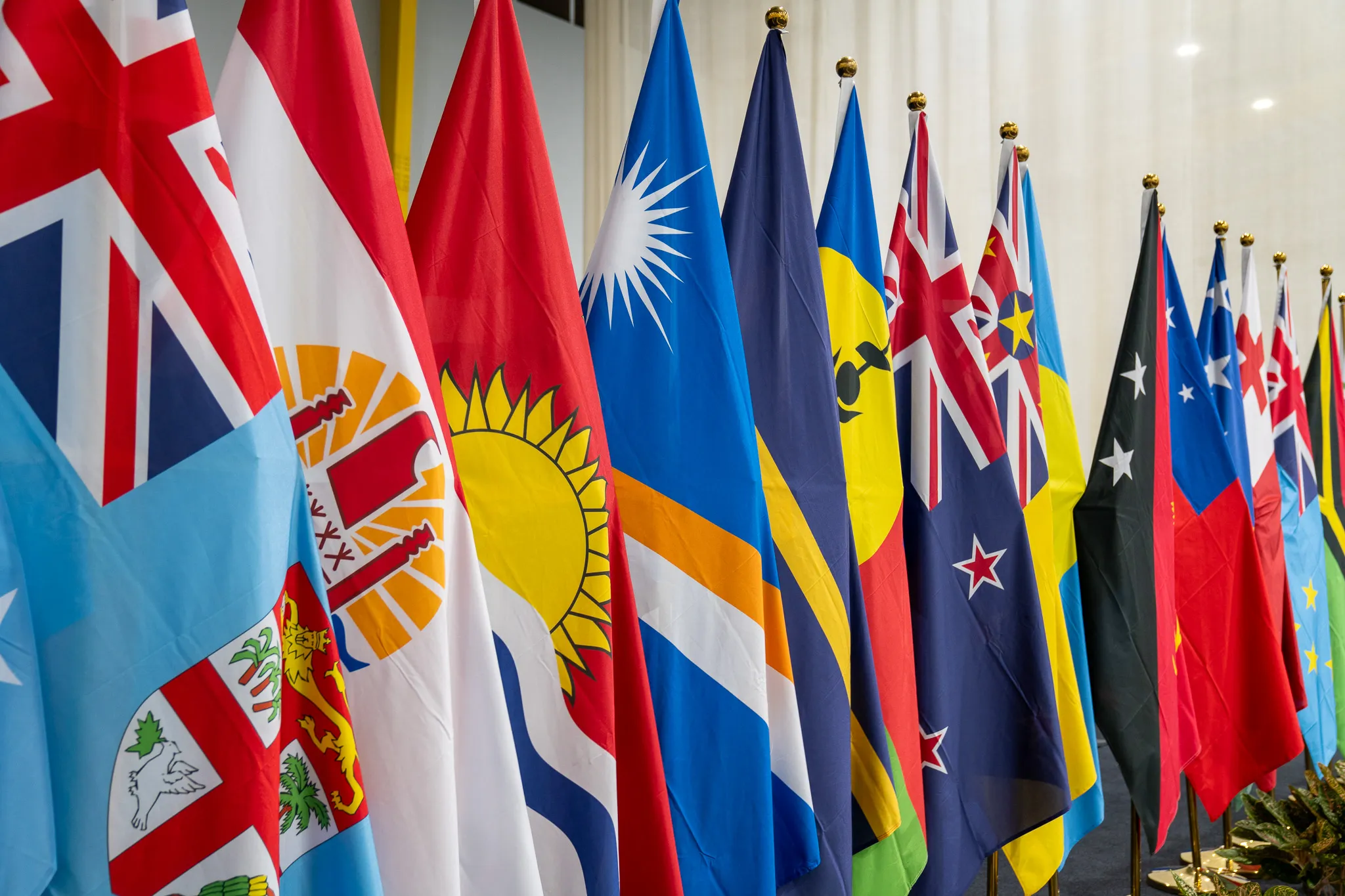 A row of colorful national flags from various countries stands indoors on flagpoles, arranged side by side at an international conference or diplomatic event. The flags feature diverse designs and colors, including blue, red, yellow, white, and black, creating a vibrant display against a neutral background.
