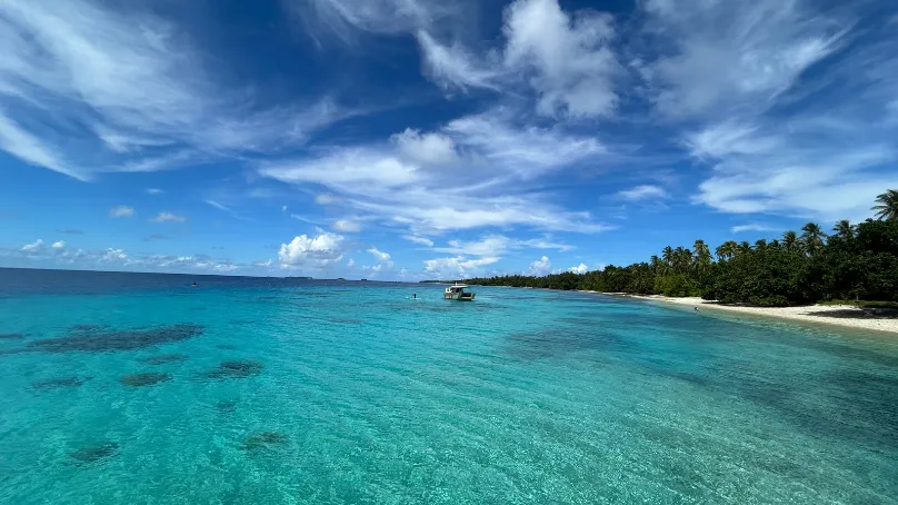 Aerial view of a tropical island coastline with clear turquoise water, a sandy beach lined with palm trees, and a small boat floating offshore beneath a bright blue sky with scattered clouds.