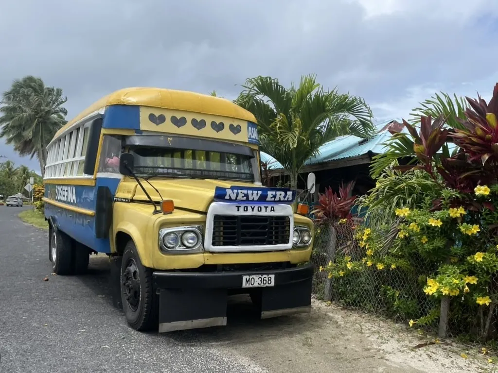 A yellow-and-blue vintage Toyota bus is parked along a quiet tropical roadside, with palm trees, colorful flowering plants, and small houses nearby. Dark gray storm clouds fill the sky above the scene.