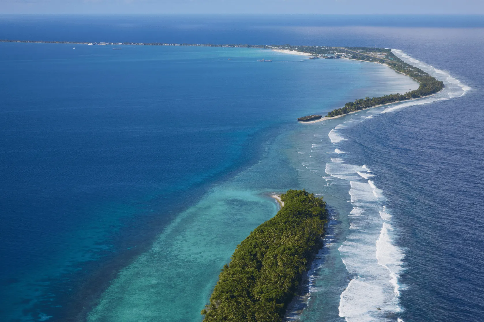 Aerial view of a Pacific island atoll showing a narrow strip of forested land surrounded by turquoise lagoon waters and ocean waves breaking along the outer reef, with a distant shoreline and scattered buildings visible in the background.